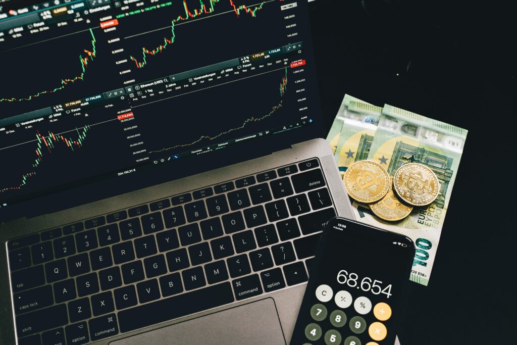 A laptop displaying stock charts with Bitcoin, Euros, and a cellphone calculator, showcasing financial analysis.